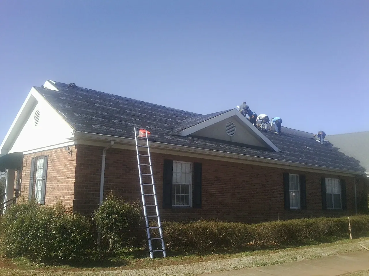 Crew tearing shingles off a brick ranch roof with a ladder set — Emergency Roof Repair work by our crew in Mc Rae Helena