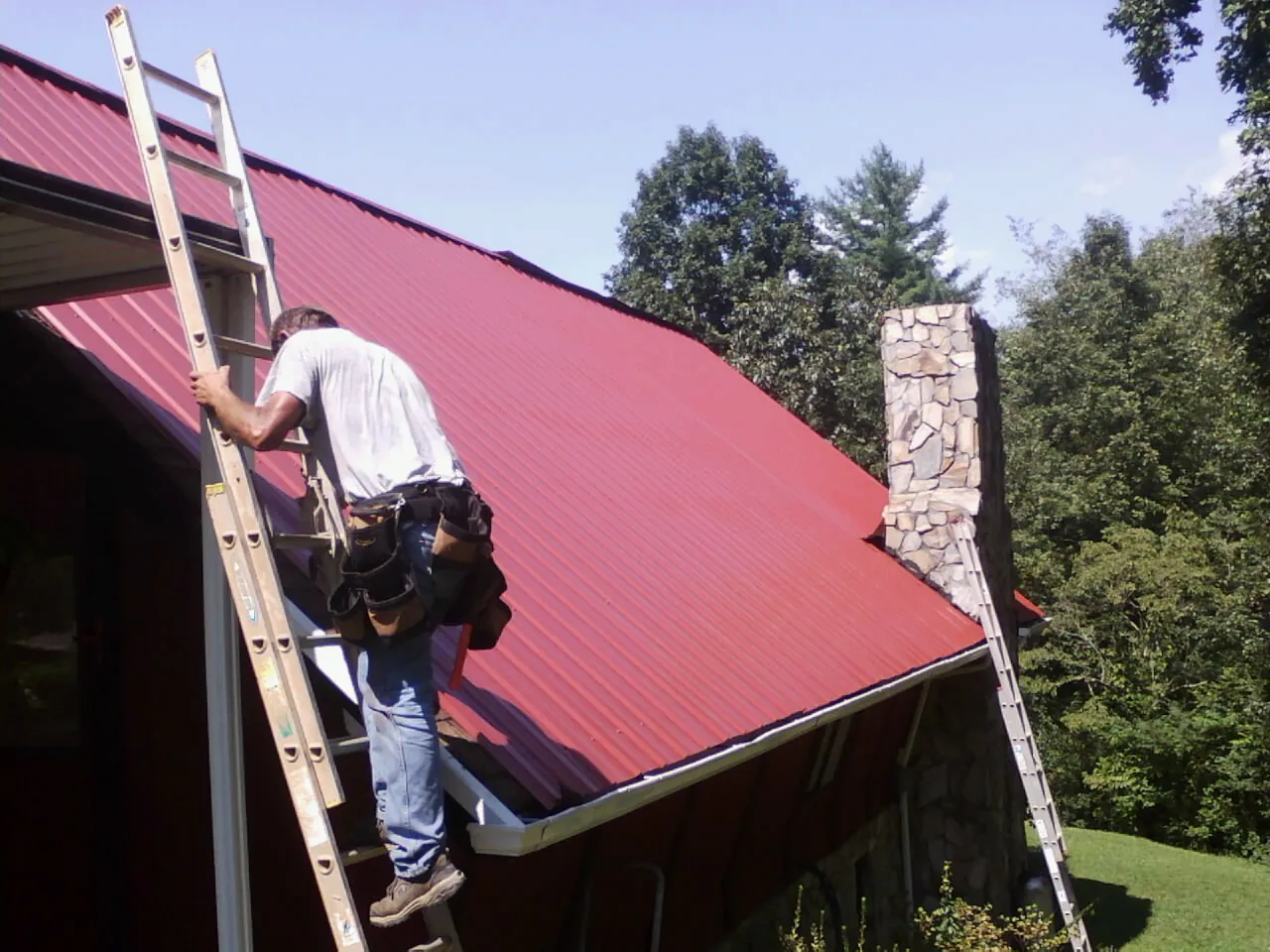 Roofer on a ladder beside a red metal roof and stone chimney — Soffit & Fascia Repair by Roofing Brothers in Mc Rae Helena